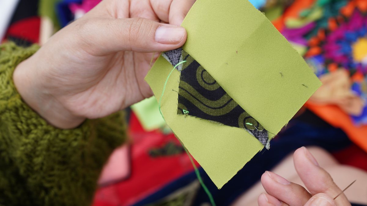 Hands piecing a green fabric square onto a colorful patchwork quilt