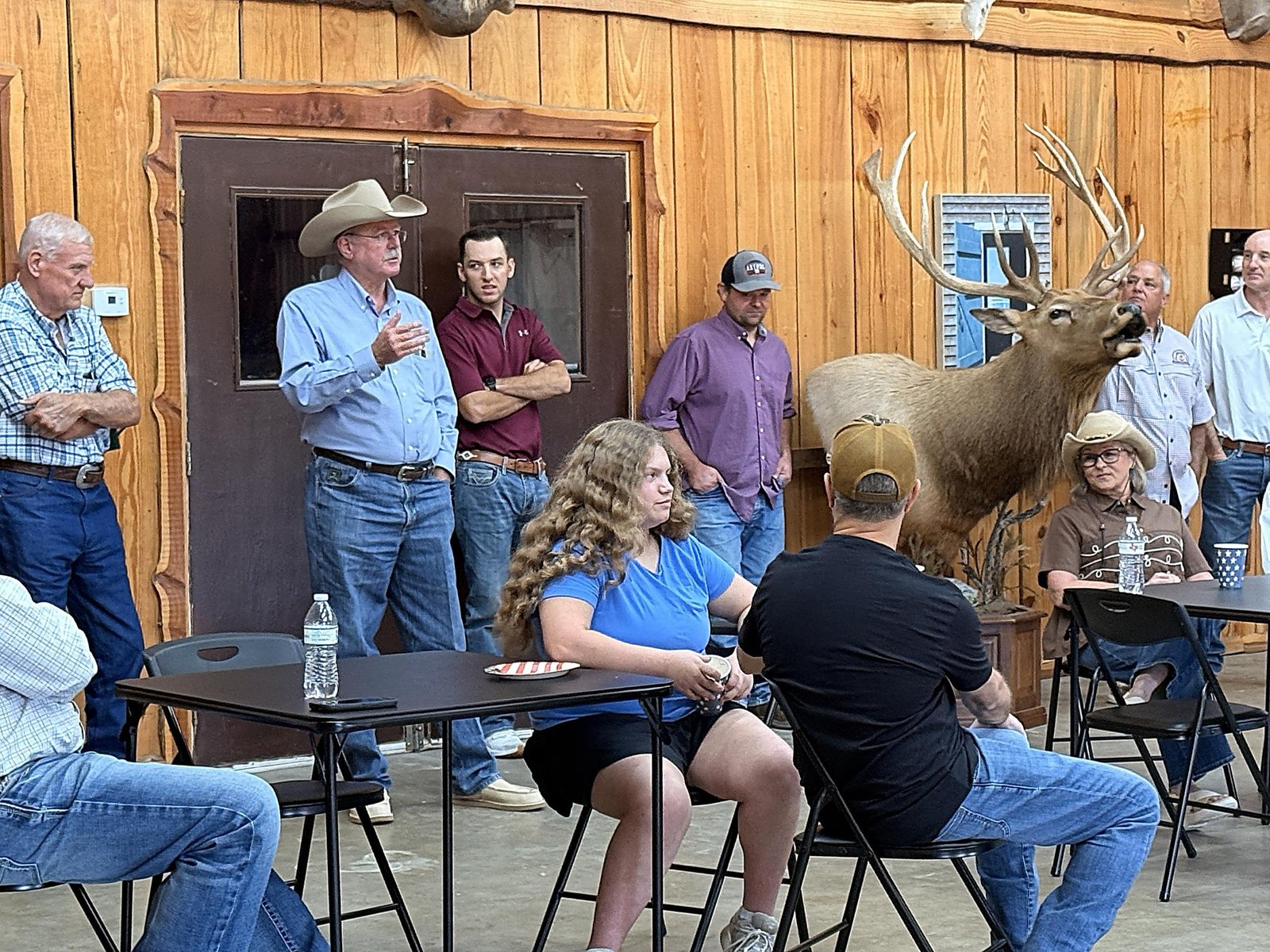 Billy Wayne Cox, in a cowboy hat, asks a question as guests sit at folding tables in a wood-paneled hall with a mounted elk.