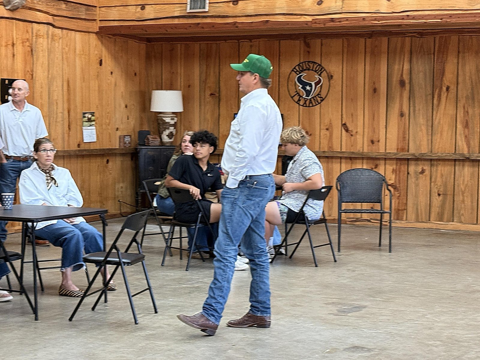 Nate Sheets, in a green cap and white shirt, stands in front of seated guests in a wood-paneled room.