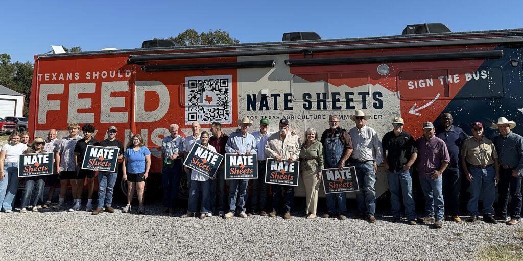 Supporters holding campaign signs stand in front of the orange and navy "Texans Should Feed Texans" campaign bus on a gravel lot.