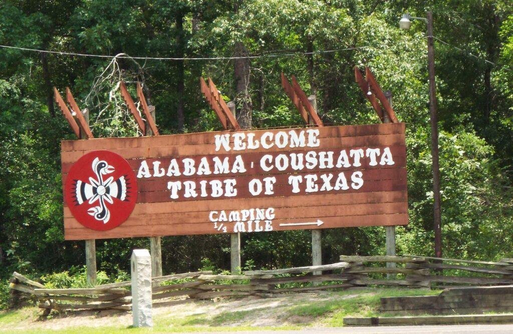 Wooden welcome sign reading Welcome Alabama-Coushatta Tribe of Texas, with the tribal seal, surrounded by pine forest