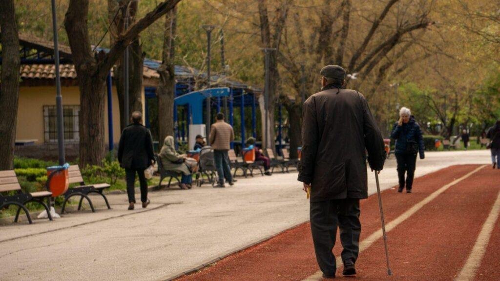 Older man walking a park path with a cane, seen from behind on a tree-lined pathway