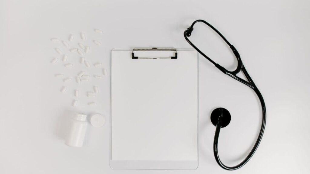 Clipboard, pill bottle and black stethoscope arranged on a white desk